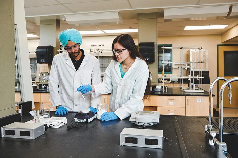 Two students working in a lab wearing white lab coats.