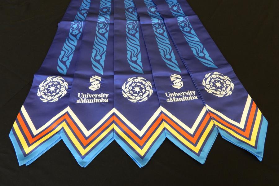 A front view of 5 purple graduation stoles with blue, yellow, red, and white chevrons. A blue patterned motif with imagery representing the sky, sun, land, flowers, water and stars runs the length of the stole. Each side of the stole is embroidered with the University of Manitoba logo, and a motif in a circular pattern.