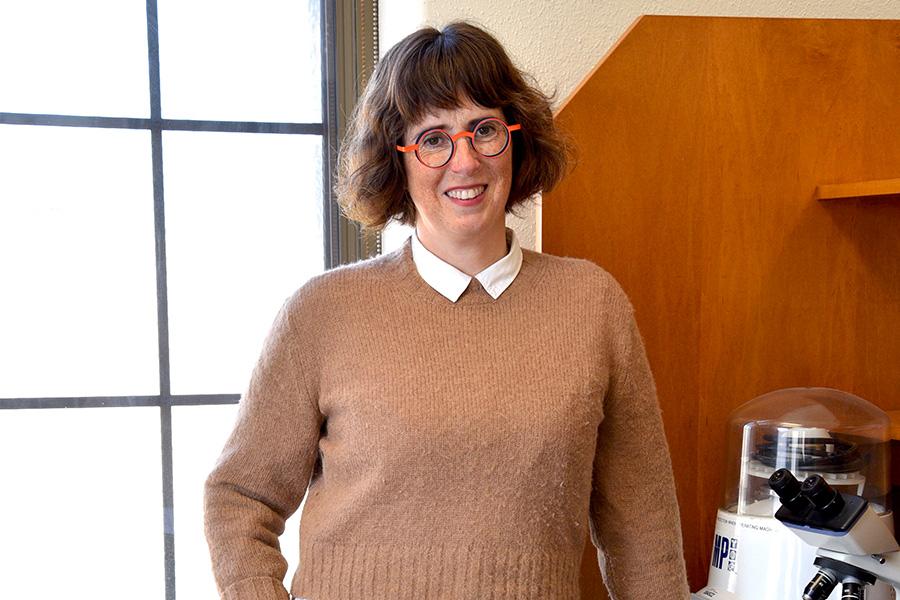 Olivia Watkins, wearing a brown sweater with white collar and round glasses stands while smiling and posing for the camera.