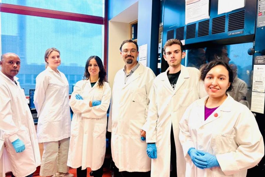 A group of researchers all wearing white lab coats with blue gloves stand in a lab smiling and posing for the camera.