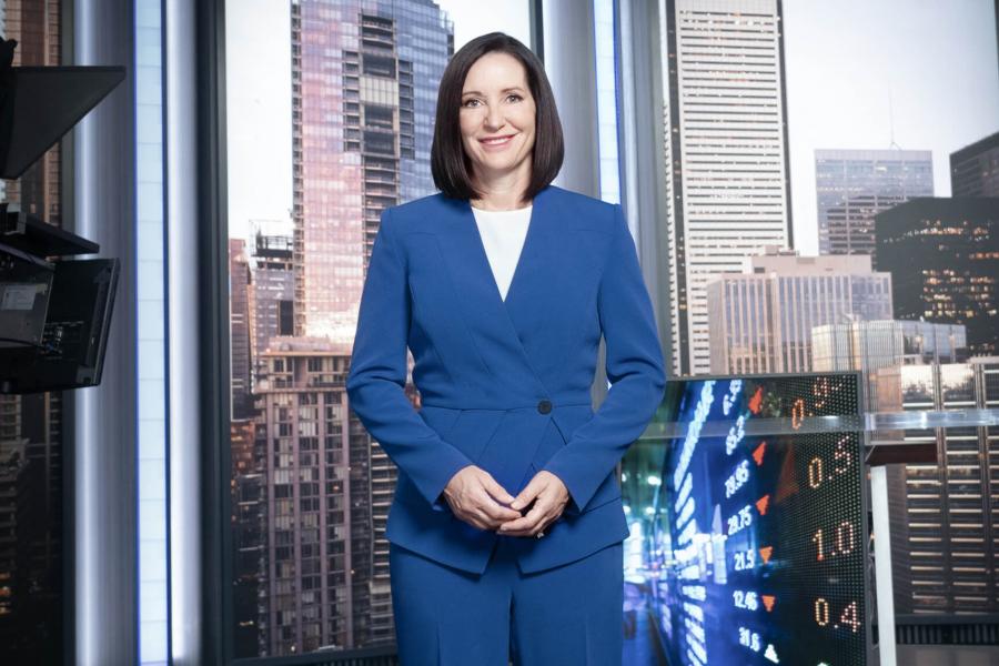 Amanda Lang stands in a blue suit with her hands together and smiling in a news studio.