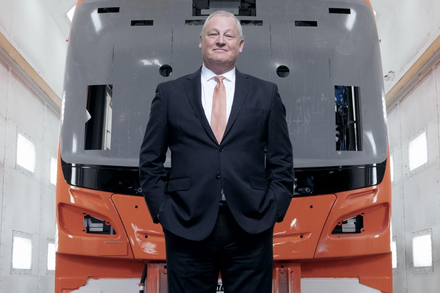 Paul Soubry in a dark suite with a white shirt and his hands in his pockets stands in front of a transit bus being built inside a well-lit garage.