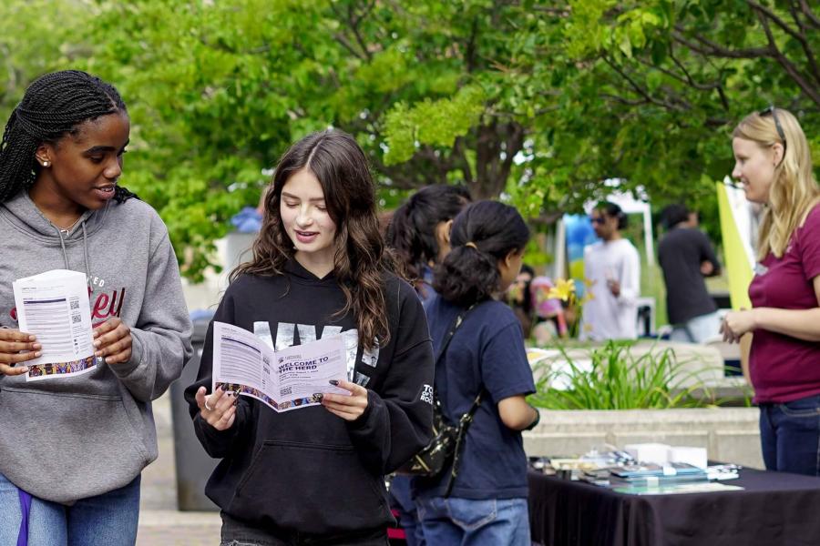 Two students walk and look at a pamphlet at an outdoor event with tables and campus tours.