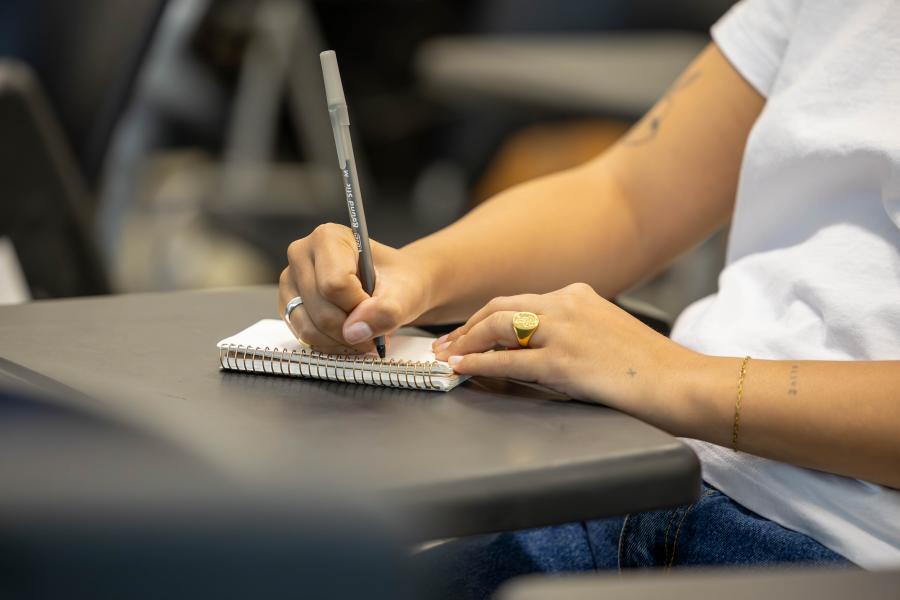 Two arms resting on a notepad sitting on a small desk. A black pen is in the right hand of the notetaker.