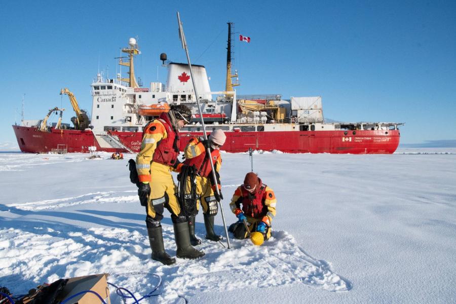 Three people pierce the ice in the arctic with the Canadian Coast Guard Ship Amundsen visible in the background.