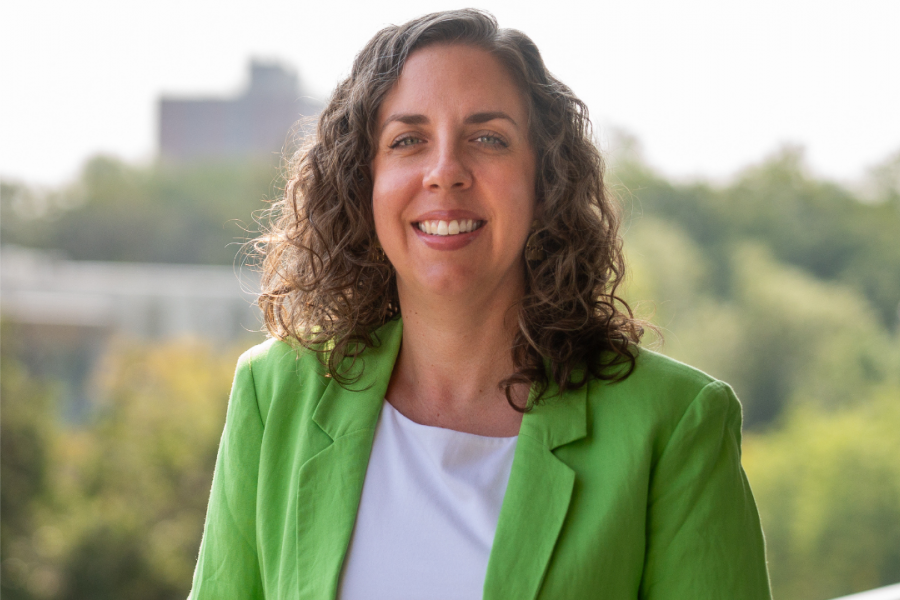 A smiling woman with curly hair stands on an outdoor balcony, wearing a bright green blazer over a white top, with trees and a blurred building in the background.