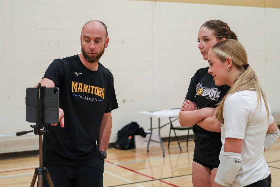 A coach points at an ipad delivering feedback to two Junior Bisons girls volleyball players.