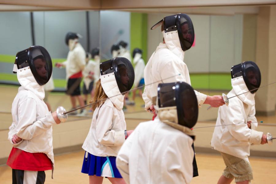 Four children wearing fencing masks and jackets practice their fencing skills guided by an instructor.