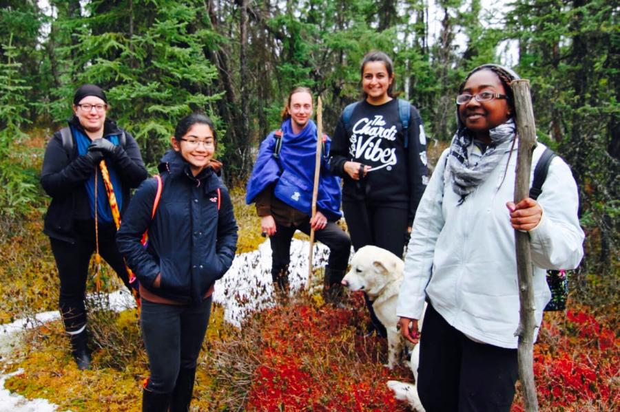 A group of five people and a dog hold walking sticks as they prepare for a hike in a wooded area.