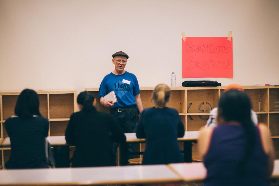 Older male standing up speaks to a group of people sitting on a table across from him.