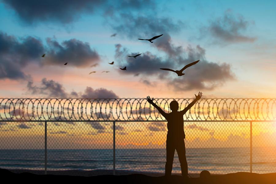 Brown Bag Lecture Series - boy with arms extended silhouetted and facing the open seas behind wire fence