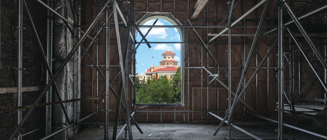 A window view from the inside of a building being constructed shows a blue sky and the Fort Garry Administration Building at UM.