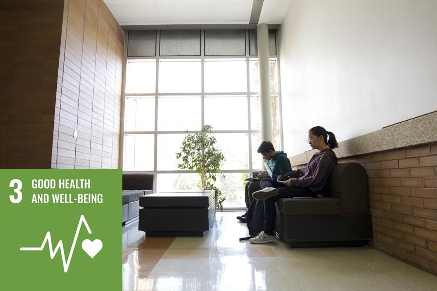 Two people sitting on a couch on the right with a bright window beyond them. A green coloured square in the corner has a logo of a heartbeat line and a heart and the text 3 GOOD HEALTH AND WELL-BEING.